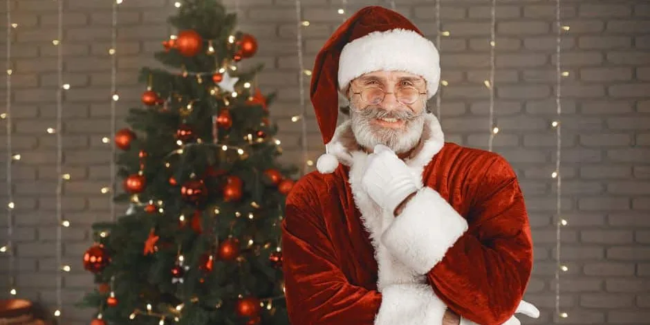 Le Père Noël souriant en costume rouge, à côté d’un sapin de Noël décoré de boules rouges et de guirlandes lumineuses (photo d’illustration)